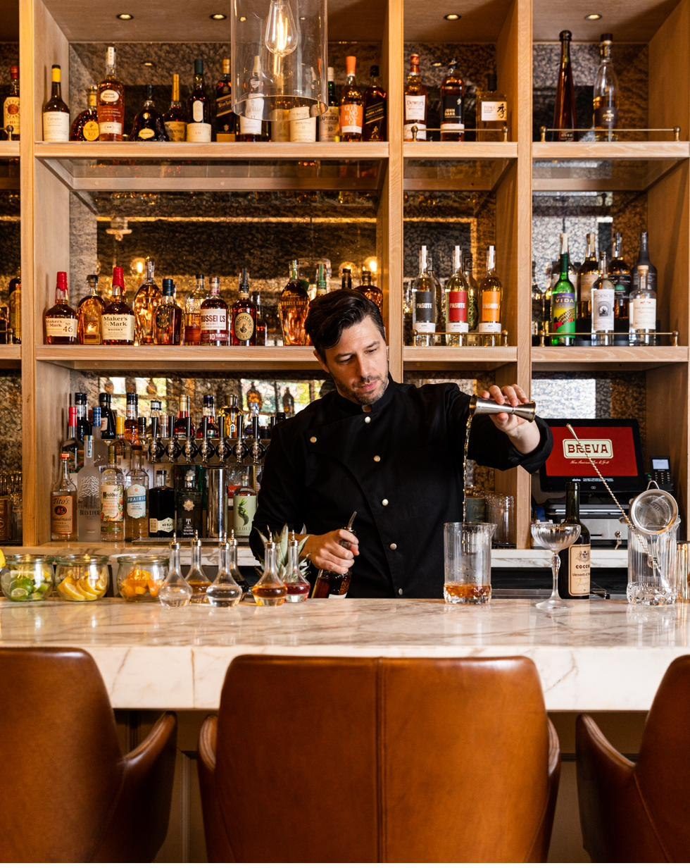 The principal behind the bar, pouring from a jigger into a mixing glass, the back bar stacked behind him.