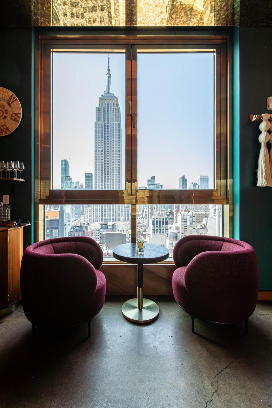 Two velvet chairs and a small table at a hotel bar window, the Empire State Building filling the glass beyond.