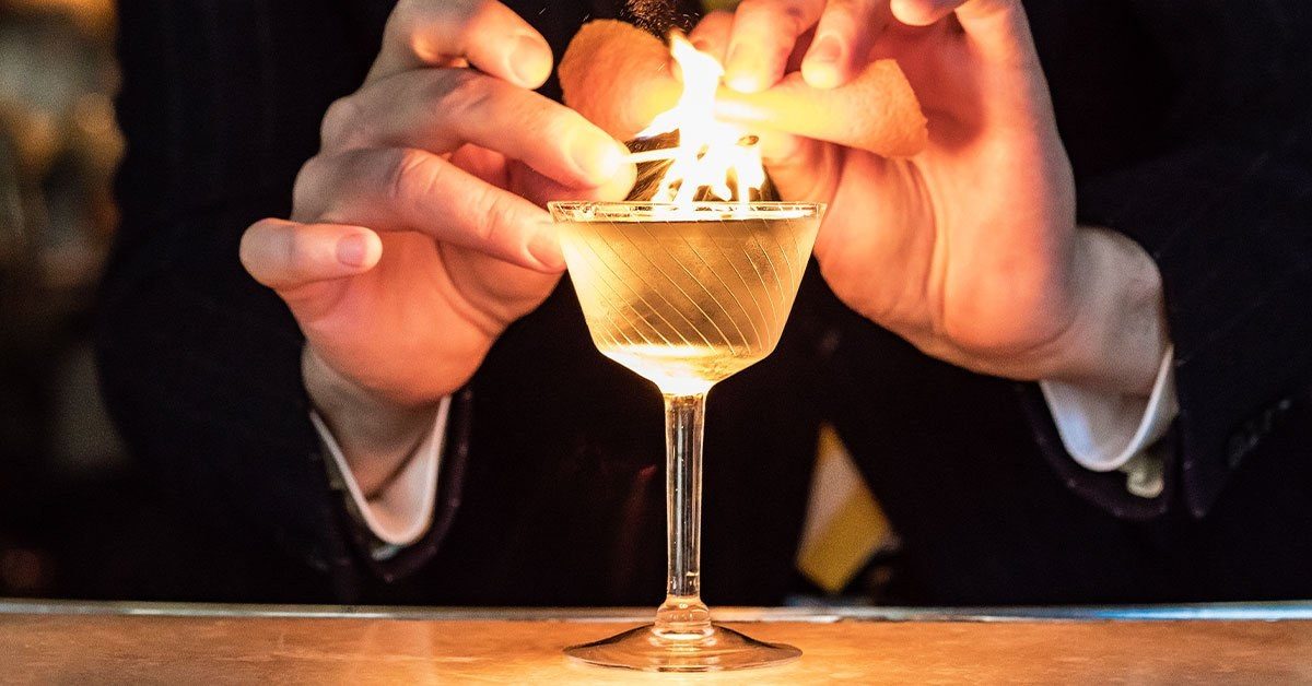 A bartender’s hands expressing a flamed citrus peel over a chilled coupe at the bar.