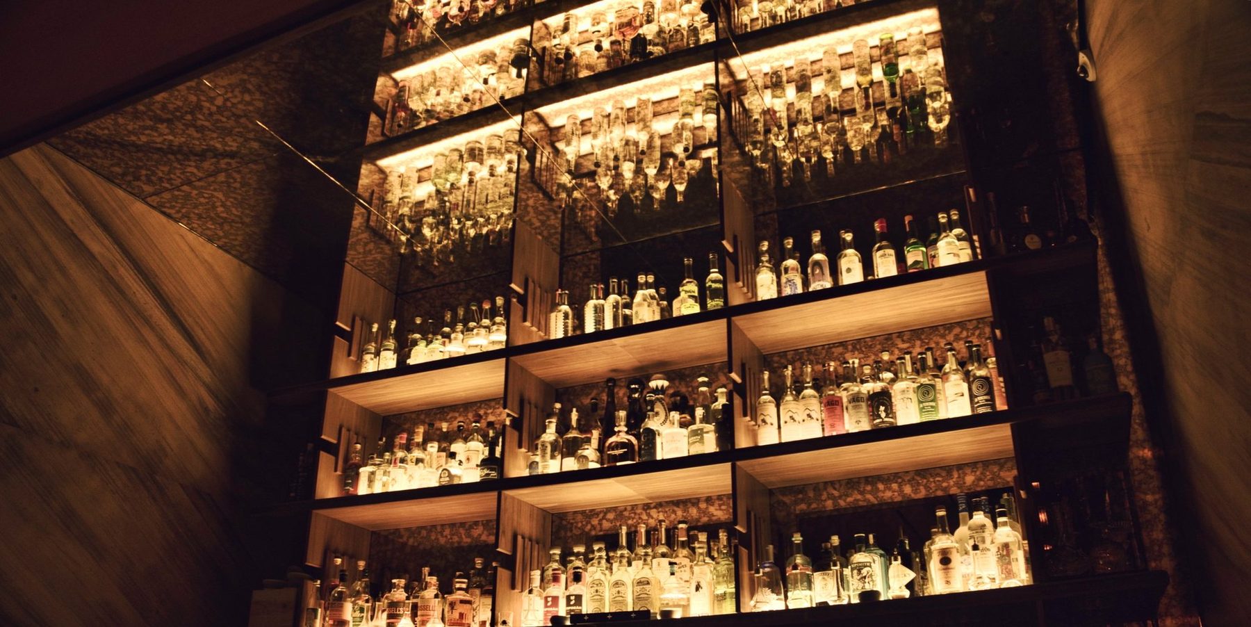 A lit back bar rising in tiers, bottles held in mirrored casework.
