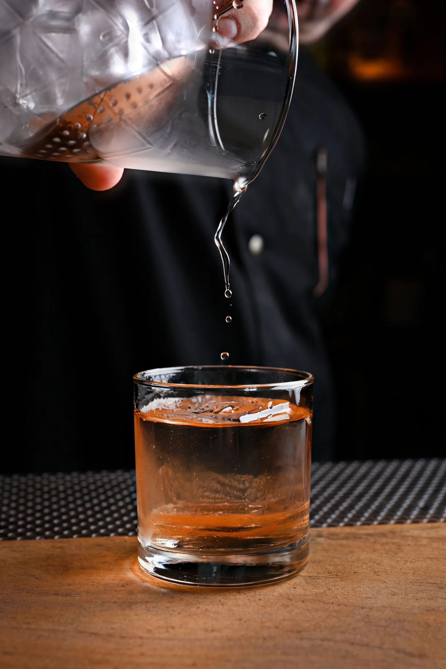 A stirred drink poured from a mixing glass into a rocks glass over one large cube, the bartender in shadow behind it.