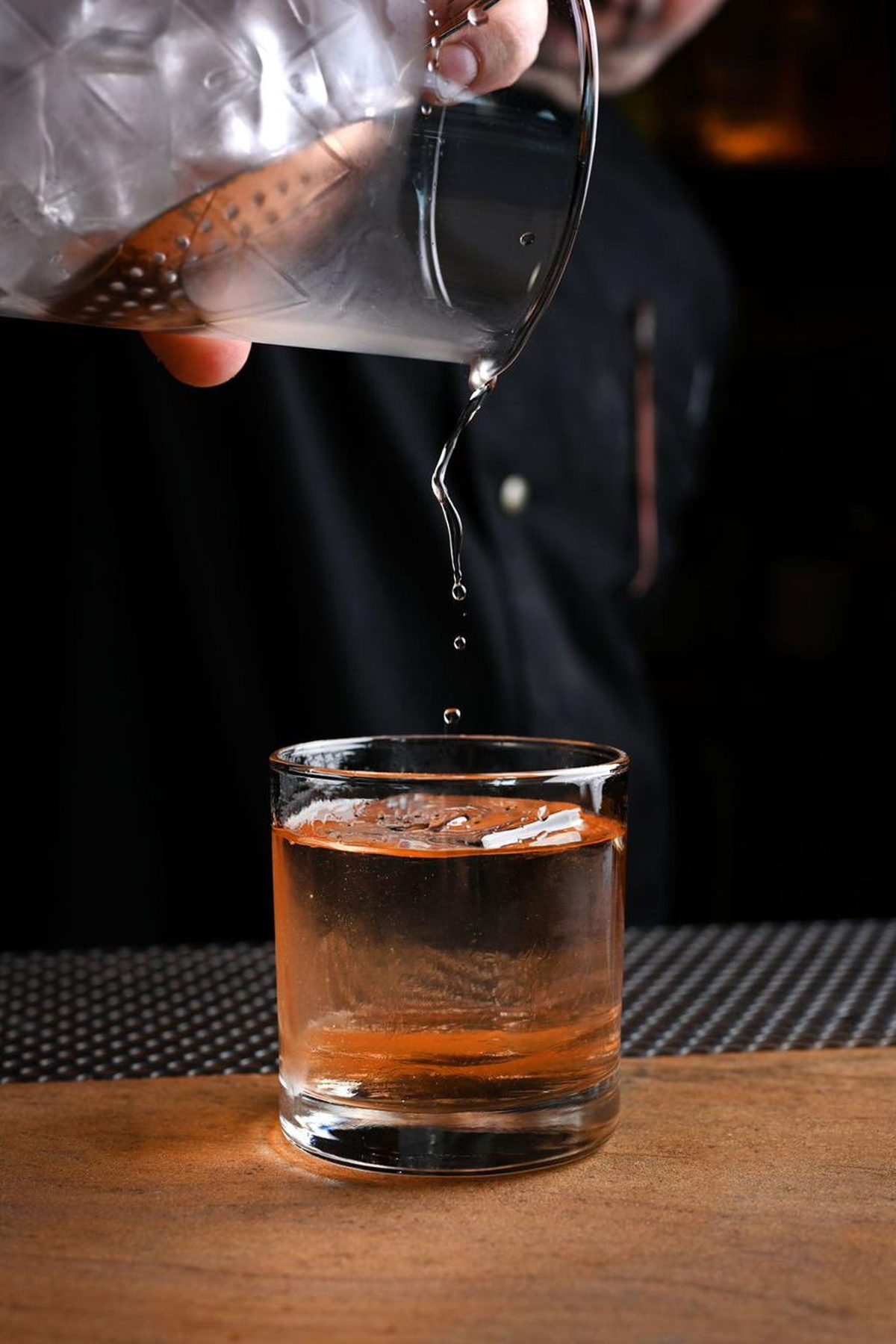 A stirred drink poured from a mixing glass into a rocks glass over one large cube, the bartender in shadow behind it.