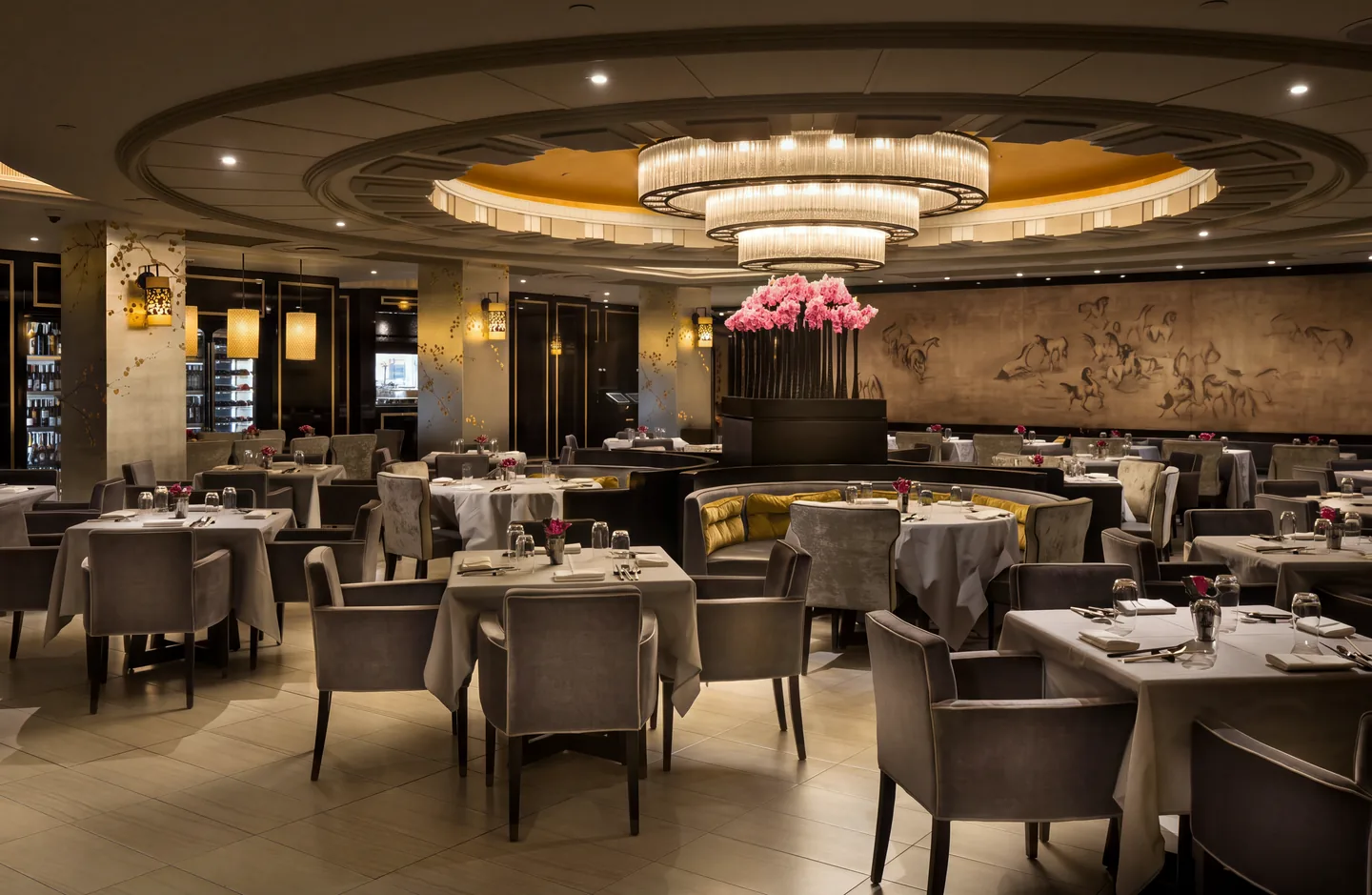 A hotel dining room laid and empty before service, lit from a single tiered chandelier, a relief mural of horses running the far wall.