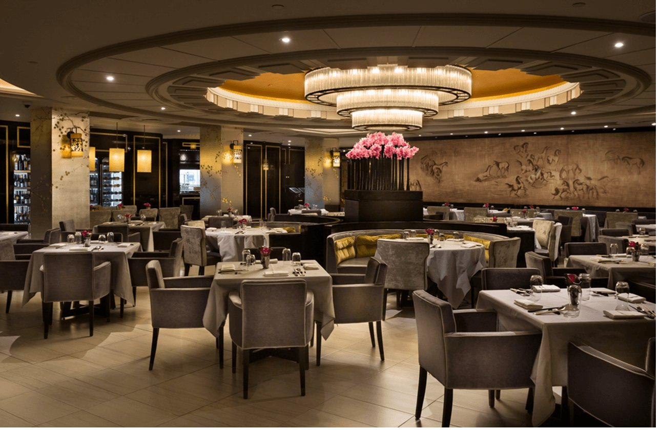 A hotel dining room laid and empty before service, lit from a single tiered chandelier, a relief mural of horses running the far wall.