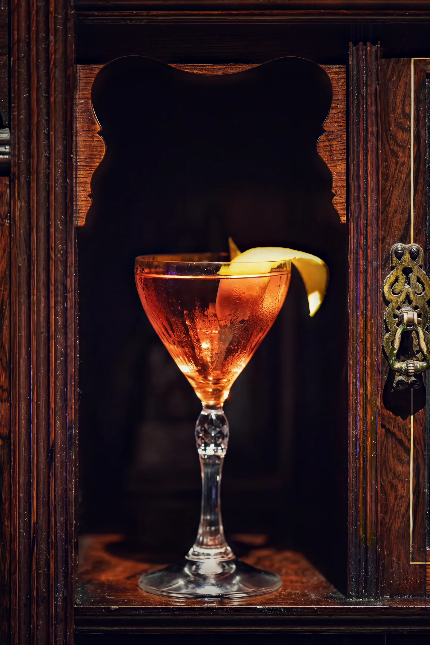 A coupe standing in a lit alcove of a carved mahogany cabinet, a lemon twist across the rim.