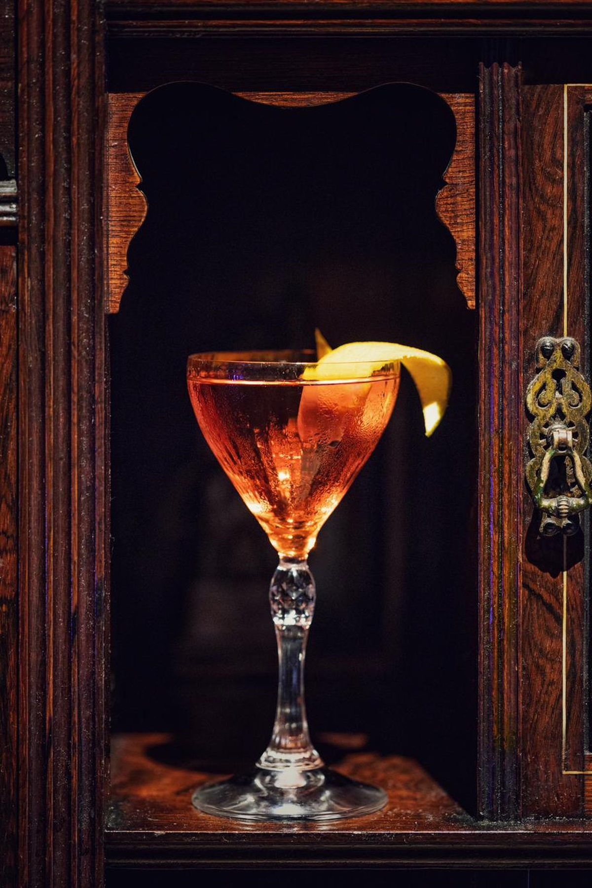 A coupe standing in a lit alcove of a carved mahogany cabinet, a lemon twist across the rim.