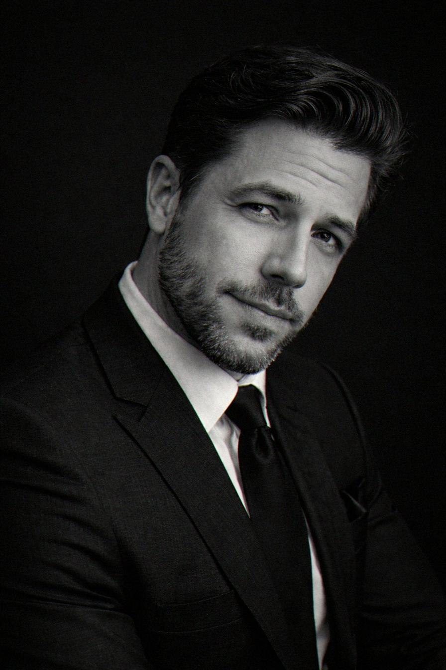 The principal in a dark suit and tie, photographed against a black ground.