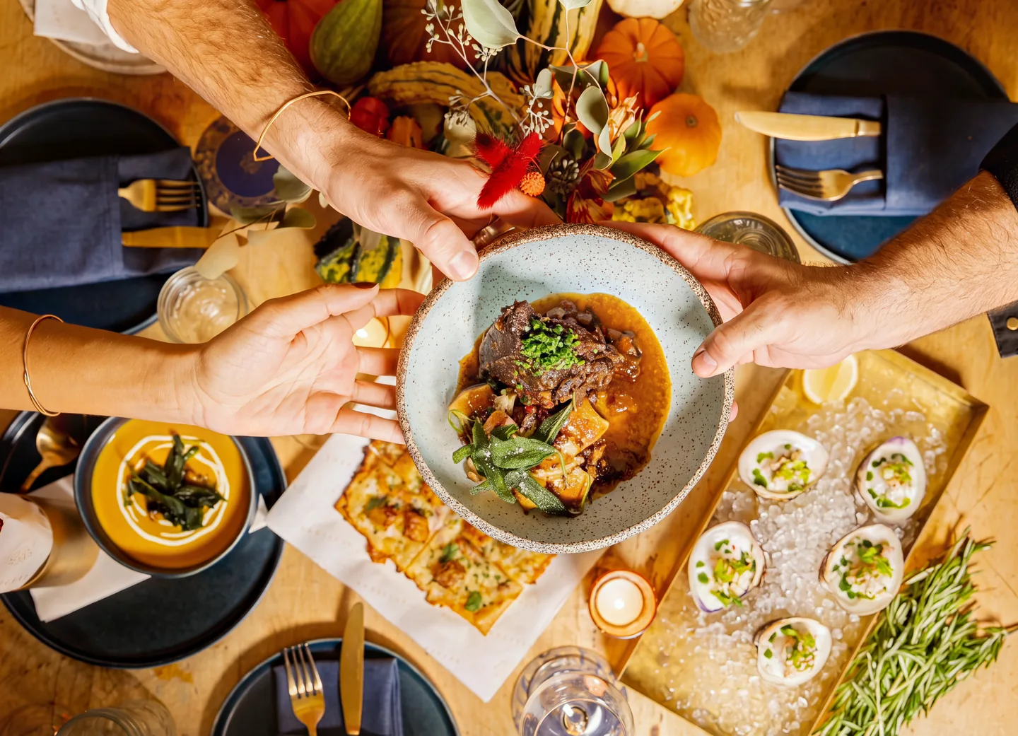A shared table seen from above, four hands passing a braise across the centre.