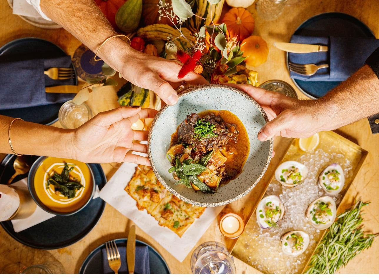 A shared table seen from above, four hands passing a braise across the centre.